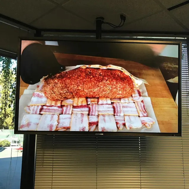 In line for lunch. A man on the tv wraps a lump of cheese in meat, then weaves a bacon jacket for it.